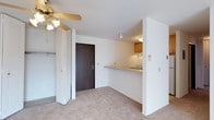 This single-floor space features an open and functional layout that connects the living area to the kitchen through a pass-through counter, creating an efficient flow between the two zones. The living area is finished with wall-to-wall carpeting in a neutral tone, contributing to a comfortable and understated ambiance. A ceiling fan with a light fixture is installed overhead, providing both ventilation and ambient lighting to the space.

A bifold closet door on one side of the room reveals a closet with built-in shelving and hanging rod, offering practical storage directly within the living area. A dark-stained wood entry door provides a visual contrast against the predominantly white walls, adding a subtle design element to the otherwise neutral palette.

The kitchen is partially separated by a half-wall with a pass-through counter, allowing visibility between the spaces while maintaining defined zones. Warm wood-toned upper cabinetry lines the kitchen walls, and under-cabinet lighting illuminates the work surface below. A stovetop is visible within the kitchen, and ceiling-mounted lighting ensures the cooking area is well-lit. The overall design is clean and practical, with a cohesive neutral color scheme throughout.