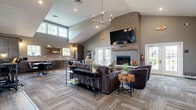 Located at 9060 Heirloom Ln in Reynoldsburg, Ohio, this well-appointed single-floor community space offers a thoughtfully designed layout that blends comfort and functionality. The centerpiece of the space is a grand living area anchored by a striking brick fireplace with a wall-mounted television above, framed by vaulted ceilings that lend an impressive sense of height and openness. Light wood-look flooring flows through the main corridor and dining area, while patterned carpet adds texture and warmth to the lounge zones. The living area is furnished with plush seating, leather accent chairs, and open metal shelving displays, creating an inviting and curated atmosphere. A dining area features a solid wood table with upholstered seating positioned beneath high windows that draw in natural light and offer views of the surrounding landscape. Glass-panel French doors open to a private office space equipped with a workstation and professional seating, maintaining visual connectivity while ensuring privacy. Adjacent to the main corridor, a fitness area is visible through floor-to-ceiling glass panels, housing cardio equipment in a clean, dedicated zone. A service counter with dark cabinetry provides a functional station near the entry. Throughout, the design balances modern industrial elements with warm, rustic touches for a welcoming communal environment.