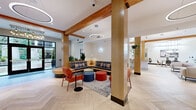 This thoughtfully designed single-floor amenity space offers a cohesive blend of warm natural materials and contemporary finishes throughout. The entry lobby welcomes visitors with a striking chandelier, built-in curved bench seating with plaid cushions, and large glass doors that flood the space with natural light. Exposed wood beams run throughout, complementing the light-toned herringbone hardwood flooring that unifies every area.

The central lounge features an L-shaped sectional sofa, colorful ottomans, and accent chairs arranged around a patterned area rug, creating a relaxed yet functional gathering space. Adjacent, a row of glass-enclosed private work pods — each distinguished by bold accent colors including green, blue, orange, and red — provides quiet, individual workstations ideal for focused tasks.

The full kitchen is equipped with warm wood cabinetry, a large central island with bar seating, a beverage station, and abundant counter space beneath a high ceiling with recessed lighting. A separate dining area features a wood table with modern chairs, pendant lighting, and framed wall art, with direct access to an outdoor pool and patio area visible through expansive black-framed windows. Lush greenery and curated artwork are found throughout, adding warmth and character to the overall space.