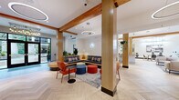This thoughtfully designed single-floor amenity space offers a cohesive blend of warm natural materials and contemporary finishes throughout. The entry lobby welcomes visitors with a striking chandelier, built-in curved bench seating with plaid cushions, and large glass doors that flood the space with natural light. Exposed wood beams run throughout, complementing the light-toned herringbone hardwood flooring that unifies every area.

The central lounge features an L-shaped sectional sofa, colorful ottomans, and accent chairs arranged around a patterned area rug, creating a relaxed yet functional gathering space. Adjacent, a row of glass-enclosed private work pods — each distinguished by bold accent colors including green, blue, orange, and red — provides quiet, individual workstations ideal for focused tasks.

The full kitchen is equipped with warm wood cabinetry, a large central island with bar seating, a beverage station, and abundant counter space beneath a high ceiling with recessed lighting. A separate dining area features a wood table with modern chairs, pendant lighting, and framed wall art, with direct access to an outdoor pool and patio area visible through expansive black-framed windows. Lush greenery and curated artwork are found throughout, adding warmth and character to the overall space.
