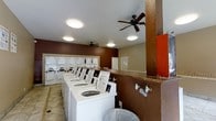 This single-floor shared laundry facility is designed to accommodate multiple users simultaneously. A row of top-loading washing machines lines one wall, each equipped with instructional signage for user guidance. Adjacent to the machines, a bank of coin-operated payment and control panels is mounted along the wall, providing a straightforward, organized system for managing laundry cycles. The room is divided by a structural column finished in a contrasting brown tone, adding a visual anchor to the open layout. The walls feature a two-tone color scheme, with deep brown on the upper portion and a lighter neutral tone along the lower half, accented by a decorative tile border. Two ceiling fans are installed overhead, promoting air circulation throughout the space and helping to manage heat and humidity generated by active machines. Recessed flush-mount light fixtures provide even, functional illumination across the room. The flooring consists of light-toned tiles, offering durability and ease of cleaning. A utility area with exposed plumbing connections is visible near the structural column, indicating provisions for additional equipment hookups. Overall, the space is utilitarian in design, prioritizing function and efficiency.