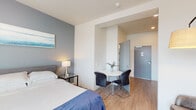 This thoughtfully designed studio unit offers a well-organized single-floor layout that maximizes functionality within a compact footprint. The sleeping area serves as the focal point of the main living space, featuring a full-sized bed with a upholstered headboard positioned against a slate-blue accent wall. Two large windows with horizontal blinds flood the room with natural light and provide views of the surrounding cityscape, enhancing the sense of openness. Light wood-tone flooring runs throughout, contributing to a clean, contemporary aesthetic.

A defined dining nook sits adjacent to the sleeping area, accommodating a round white table with two sleek black chairs—an efficient use of space for everyday dining. The compact kitchenette is outfitted with gray cabinetry, open shelving, a full-size refrigerator, a cooktop, a microwave, a sink, and a coffee maker, offering practical self-catering capabilities.

The bathroom features a white vanity with a large mirror, a shower with curtain enclosure, and light wood-tone flooring consistent with the rest of the unit. The overall design is clean and modern, with neutral walls, recessed lighting, and a cohesive color palette of whites, grays, and soft blues throughout.