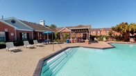 This single-story residence is part of a well-established brick exterior community that offers both comfortable interior living and inviting shared outdoor amenities. The community's outdoor area centers around a sparkling swimming pool with a brick-trimmed surround, complemented by a wooden pergola structure that provides shaded seating. Additional lounge chairs and picnic tables are arranged around the pool deck, creating a relaxed and social atmosphere under open skies.

Inside, the unit features consistent wood-look flooring throughout, lending warmth and visual continuity across the living spaces. The main living area is highlighted by a ceiling fan with integrated lighting and a pass-through window that connects directly to the kitchen, offering a practical and open feel between the two spaces. Natural light filters in through blinds-fitted windows, brightening the interior rooms.

The entryway is clean and functional, with a solid white front door and straightforward layout that leads into the living areas. A staircase is visible in one section of the interior, along with a hallway corridor that provides access to additional rooms and storage. The interior palette is neutral throughout, with white walls and clean trim work that create a bright, move-in-ready environment.