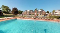 This residential community showcases a well-appointed outdoor amenity area centered around a generously sized swimming pool with clear, blue water. The pool deck is finished in a warm terracotta-toned surface and lined with multiple lounge chairs, providing ample space for relaxation and sunbathing. The area is enclosed by a wrought iron fence, offering both security and an open, airy feel.

Surrounding the pool, the community features classic multi-story brick buildings with pitched roofs, large windows, and private balconies, reflecting a traditional architectural style. A prominent brick chimney serves as a distinctive architectural focal point rising above the roofline. Lush, mature trees are scattered throughout the property, adding greenery and shade to the overall environment.

The grounds are well-maintained, with open parking areas and walkways connecting the various buildings. The community benefits from abundant natural light, as evidenced by clear blue skies overhead, creating a bright and inviting outdoor atmosphere. The combination of the sparkling pool, classic brick architecture, and landscaped surroundings gives the property a comfortable, established character well-suited for everyday residential living.