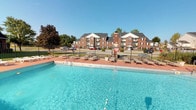 This residential community showcases a well-appointed outdoor amenity area centered around a generously sized swimming pool with clear, blue water. The pool deck is finished in a warm terracotta-toned surface and lined with multiple lounge chairs, providing ample space for relaxation and sunbathing. The area is enclosed by a wrought iron fence, offering both security and an open, airy feel.

Surrounding the pool, the community features classic multi-story brick buildings with pitched roofs, large windows, and private balconies, reflecting a traditional architectural style. A prominent brick chimney serves as a distinctive architectural focal point rising above the roofline. Lush, mature trees are scattered throughout the property, adding greenery and shade to the overall environment.

The grounds are well-maintained, with open parking areas and walkways connecting the various buildings. The community benefits from abundant natural light, as evidenced by clear blue skies overhead, creating a bright and inviting outdoor atmosphere. The combination of the sparkling pool, classic brick architecture, and landscaped surroundings gives the property a comfortable, established character well-suited for everyday residential living.