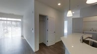 This single-floor space showcases a contemporary design with a clean, neutral aesthetic throughout. The interior is finished with dark hardwood-style flooring that contrasts sharply against the crisp white walls and white baseboards, creating a polished, modern look. Recessed lighting is installed in the ceiling, supplemented by a stylish pendant light with a brushed gold fixture and white shade, visible near what appears to be a kitchen counter area with a bright white quartz or solid-surface countertop edge. The walls are painted a uniform white, contributing to a bright and open atmosphere. A paneled white door opens to reveal a closet with a hanging rod, offering practical built-in storage. The layout suggests an open-concept flow between spaces, with a hallway extending toward additional rooms. Natural light filters in from what appears to be a window further down the corridor, enhancing the overall brightness of the space. The design choices throughout — from the hardware finishes to the flooring — reflect a cohesive, move-in-ready aesthetic suited to modern living.