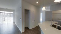 This single-floor space showcases a contemporary design with a clean, neutral aesthetic throughout. The interior is finished with dark hardwood-style flooring that contrasts sharply against the crisp white walls and white baseboards, creating a polished, modern look. Recessed lighting is installed in the ceiling, supplemented by a stylish pendant light with a brushed gold fixture and white shade, visible near what appears to be a kitchen counter area with a bright white quartz or solid-surface countertop edge. The walls are painted a uniform white, contributing to a bright and open atmosphere. A paneled white door opens to reveal a closet with a hanging rod, offering practical built-in storage. The layout suggests an open-concept flow between spaces, with a hallway extending toward additional rooms. Natural light filters in from what appears to be a window further down the corridor, enhancing the overall brightness of the space. The design choices throughout — from the hardware finishes to the flooring — reflect a cohesive, move-in-ready aesthetic suited to modern living.