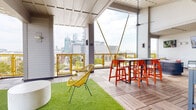 Located in the South End neighborhood of Charlotte, NC, this rooftop amenity level offers a dynamic blend of indoor and outdoor spaces designed for both relaxation and socializing. The open-air deck is finished with wood-tile decking and features distinct zones for lounging and dining. A lush artificial turf area anchors the lounge section, furnished with vibrant yellow woven chairs and a cylindrical concrete ottoman, while a bar-height dining table with bold orange stools provides a dedicated space for gathering. A built-in hot tub sits along the perimeter of the deck, accompanied by an outdoor sectional sofa and a wall-mounted television, creating a comfortable outdoor living environment. Gold-toned cable railings frame the deck on multiple sides, offering unobstructed panoramic views of the Charlotte skyline. A striking geometric gold-accented glass partition serves as both a design focal point and a wind barrier. The indoor portion of the space features soaring ceilings, walls in a soft mint green, and distinctive hexagonal floor-to-ceiling tile wainscoting in deep charcoal. Large windows flood the room with natural light and maintain visual connection to the outdoor skyline. A conference-style table with upholstered chairs and a wall-mounted television make this area well-suited for meetings or private events.