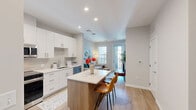 Located at 303 Lincoln St in Marlborough, MA, this well-appointed 2-bedroom, 1-bathroom residence offers a thoughtfully designed single-floor layout with modern finishes throughout. The open-concept kitchen is a standout feature, showcasing crisp white cabinetry, stainless steel appliances including a range, microwave, refrigerator, and dishwasher, complemented by a tile backsplash and light wood-look flooring. A central island with seating provides an ideal casual dining area. The living room is bright and welcoming, with large windows and a glass door opening directly onto a private balcony, offering views of the well-maintained courtyard below. The primary bedroom is generously sized, featuring hardwood-style flooring, a large window for natural light, and a spacious walk-in closet with wire shelving. The second bedroom also benefits from a bright window with exterior views. The bathroom is cleanly finished with a walk-in glass shower, wood-toned vanity, and wood-look tile flooring. Building amenities include a landscaped communal courtyard with seating, fire pit, and lawn areas. The modern exterior features a mix of gray and red paneling with private balconies throughout.