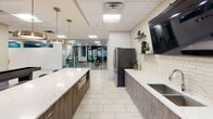 Located at 115 Second Ave S in Minneapolis, MN, this single-floor amenity space combines modern design with practical functionality. Light-toned tile flooring flows throughout, unifying each distinct area under a suspended grid ceiling with recessed panel lighting.

The lounge area serves as a central gathering hub, anchored by a generous sectional sofa in charcoal gray accented with teal throw pillows, a wooden coffee table with integrated power outlets, and a vibrant orange accent chair. Bold, colorful artwork adorns a deep navy feature wall, adding an artistic focal point to the relaxed seating environment. Direct access to an outdoor courtyard is provided through large dark-framed sliding glass doors.

The kitchen is outfitted with sleek gray cabinetry, quartz countertops, a stainless steel refrigerator, and a wall-mounted television above subway tile backsplash. Pendant lighting and a large island make this a well-appointed communal space. Windows overlooking the courtyard bring in natural light.

A glass-walled conference room offers transparency and professionalism, furnished with a round table, modern chairs, and vibrant artwork, with outdoor views reinforcing an open atmosphere. An adjacent glass-enclosed corridor with colorful art installations adds a gallery-like quality.

The fitness center, visible through glass partitions, is equipped with strength and cardio machines. A dual water fountain station rounds out the wellness amenities, reflecting a thoughtfully curated, full-service environment.