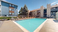 Located at 243 W 80th Ave in Denver, Colorado, this outdoor pool area serves as a central community amenity within a multi-building residential complex. The centerpiece is a generously sized rectangular swimming pool with clear, blue-green water, accessible via stainless steel entry rails. The surrounding concrete deck provides ample space for relaxation and is lined with multiple lounge chairs and chaise seating, ideal for sunbathing under Colorado's abundant sunshine. A covered pergola area with additional seating, including a woven hanging chair, offers a shaded retreat alongside the pool deck. The pool area is enclosed by a sturdy metal security fence, ensuring both safety and privacy, with manicured greenery and mature evergreen trees adding natural character to the setting. A dedicated single-story pool house with a pitched roof sits adjacent to the pool, housing utility and changing facilities. Both the American and Colorado state flags are displayed prominently on a flagpole nearby. The surrounding multi-story residential buildings, featuring a mix of stone, siding, and modern accent panels, frame the space and contribute to a cohesive community atmosphere. The overall environment is open, sun-filled, and well-maintained, offering residents a welcoming outdoor retreat.