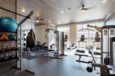 This fitness center is organized into distinct functional zones, offering a comprehensive workout environment on a single level. Entry is through glass-panel doors into a lobby-style foyer featuring a hydration station with a bottle-fill fountain and a hand sanitizer dispenser, providing a clean and practical transition into the facility.

The main gym floor is finished with durable rubber flooring and features an impressive wood-paneled ceiling with ceiling fans and recessed lighting, lending warmth and texture to the space. Large windows along one wall admit generous natural light and provide outdoor views, enhancing the open, airy atmosphere. This primary area houses Smith machines with Olympic plates, a cable system, adjustable benches, a heavy bag, kettlebells, medicine balls, and a full rack of dumbbells, supported by floor-to-ceiling mirrors that maximize both light and sightlines.

A separate studio room features light hardwood flooring, mirrored walls, a barre rail, a mounted flat-screen TV set within a distinctive geometric white accent wall, and a plyo box, making it well-suited for stretching, yoga, or small group training. The facility is thoughtfully designed to accommodate a broad range of fitness activities in a modern, well-appointed setting.