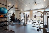 This fitness center is organized into distinct functional zones, offering a comprehensive workout environment on a single level. Entry is through glass-panel doors into a lobby-style foyer featuring a hydration station with a bottle-fill fountain and a hand sanitizer dispenser, providing a clean and practical transition into the facility.

The main gym floor is finished with durable rubber flooring and features an impressive wood-paneled ceiling with ceiling fans and recessed lighting, lending warmth and texture to the space. Large windows along one wall admit generous natural light and provide outdoor views, enhancing the open, airy atmosphere. This primary area houses Smith machines with Olympic plates, a cable system, adjustable benches, a heavy bag, kettlebells, medicine balls, and a full rack of dumbbells, supported by floor-to-ceiling mirrors that maximize both light and sightlines.

A separate studio room features light hardwood flooring, mirrored walls, a barre rail, a mounted flat-screen TV set within a distinctive geometric white accent wall, and a plyo box, making it well-suited for stretching, yoga, or small group training. The facility is thoughtfully designed to accommodate a broad range of fitness activities in a modern, well-appointed setting.