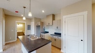 Located in Holland, Michigan, this single-floor residence offers a thoughtfully designed open-concept layout with clean, modern finishes throughout. The kitchen serves as the heart of the space, anchored by a central island with a double-basin sink and dark countertops that contrast elegantly with the light-toned cabinetry. Pendant lighting hangs above the island, providing both task illumination and a contemporary design touch. Ample upper and lower cabinetry lines the kitchen walls, offering generous storage, while a dedicated space accommodates a refrigerator. Recessed lighting and light wood-style flooring extend through the main living area, contributing to a bright and cohesive atmosphere. A dedicated nook within the kitchen provides additional counter space and storage versatility. The bedroom is finished with plush carpeting and features a wide window that frames sweeping views of open fields and autumn foliage, filling the room with natural light and a tranquil, rural backdrop. The bathroom is clean and functional, equipped with a single-sink vanity with white countertops, a toilet, and a tub-shower combination. Warm neutral tones carry throughout the entire unit, creating a calm and inviting ambiance well-suited to everyday living.