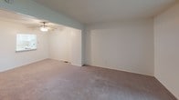 This three-floor residence offers a practical and versatile layout across its levels. The main living area is carpeted and generously sized, featuring an open passthrough to an adjoining room fitted with a ceiling fan, creating a connected and airy feel. A staircase with a wood banister leads to the upper floor, where additional carpeted bedrooms provide comfortable, quiet retreats. One bedroom benefits from a sliding glass door that opens to an outdoor balcony, offering views of surrounding greenery and natural light. A second bedroom features a standard window with exterior views. The kitchen is efficiently designed in an L-shaped configuration, equipped with white raised-panel cabinetry, light countertops, a coil-burner electric range, and a built-in overhead ventilation hood. The full bathroom includes a single-sink vanity with a mirrored cabinet, a tiled shower-tub combination, and tile flooring. The lower level consists of an expansive unfinished basement with a painted concrete floor, exposed wood-beam ceiling, and utility access, providing substantial storage or potential for future finishing. Throughout, the interior maintains a neutral, clean palette with textured ceilings on the upper levels.