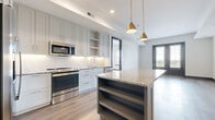 This contemporary single-floor residence showcases a thoughtfully designed open-concept layout that seamlessly connects the kitchen and living areas. The kitchen is a standout feature, equipped with stainless steel appliances including a range, microwave, dishwasher, and refrigerator. Crisp white shaker-style cabinetry extends to the ceiling, complemented by a white subway tile backsplash and granite countertops. A center island with open shelving below and a matching granite surface provides additional prep space and storage, while gold pendant lights add a refined touch overhead. Warm hardwood flooring flows throughout the main living areas, enhancing the bright, airy atmosphere created by recessed lighting and natural light from large windows and black-framed French doors that open to an outdoor balcony. The bedroom features plush neutral carpet, a ceiling fan, and a window with an outdoor view. The bathroom is well-appointed with gray shaker vanity cabinetry, granite countertops, a framed mirror with wall sconces, tile flooring, and a full tub-shower combo with white tile surround. The overall design balances modern finishes with a clean, neutral palette throughout.