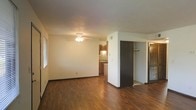 This single-floor space offers a practical and comfortable layout designed for efficient living. The main living area is anchored by rich, dark hardwood-style flooring that runs throughout, creating a warm and cohesive aesthetic. A ceiling fan with a light fixture provides both illumination and air circulation, adding to the room's functionality. Large windows allow natural light to filter in, brightening the open space. A partial wall divides the living area from the storage and entry zones, lending a sense of structure without closing off the floor plan. Closet space is generously provided, with what appears to be both a standard reach-in closet and an additional storage area fitted with louvered doors, offering ample organizational options. The kitchen, visible through an open passageway, features warm wood cabinetry, a decorative tile backsplash, and under-cabinet lighting that highlights the counter workspace. The neutral white walls throughout serve as a clean backdrop, allowing the darker flooring and cabinetry tones to stand out. Overall, the space presents a tidy, functional environment with a warm, understated character.