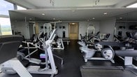 This fitness center offers a comprehensive workout environment thoughtfully arranged across a single floor. The space is outfitted with an impressive selection of cardio equipment, including elliptical trainers and recumbent bikes, as well as dedicated strength training machines to support a full-body exercise routine. The dark rubber flooring provides a durable, slip-resistant surface well-suited for high-intensity activity, while floor-to-ceiling mirrored walls along one side visually expand the space and allow users to monitor their form during workouts. Recessed track lighting overhead ensures bright, even illumination throughout the facility. A wide corridor visible toward the rear of the space suggests additional rooms or amenities beyond the main workout floor, adding to the overall functionality of the facility. The clean, modern aesthetic — with a neutral grey and white color palette — creates a focused and professional atmosphere. The layout is open and well-organized, allowing for comfortable movement between equipment. Overall, this fitness center is designed to deliver a well-rounded exercise experience in a practical and efficiently utilized space.