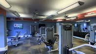 This single-level fitness center is designed to support a comprehensive strength training experience. The space is outfitted with multiple Precor cable and resistance machines arranged in an open floor layout, allowing for comfortable movement between stations. The flooring consists of patterned carpet tiles, providing a durable and practical surface suited to a gym environment. The walls are painted in a deep navy blue, accented by a bold red band running along the upper portion, lending the space an energetic and motivating atmosphere. Multiple ceiling fans are suspended throughout, ensuring adequate air circulation during workouts. Overhead tube lighting supplements the ambient light, keeping the space evenly illuminated. A wall-mounted flat-screen television is positioned to be visible from multiple workout stations, adding an entertainment element to the training area. Additional equipment is visible toward the rear of the space, suggesting further workout options beyond the primary strength machines. The overall design is functional and purposeful, with a layout that maximizes equipment accessibility while maintaining clear sightlines throughout the room.