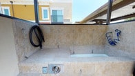 This outdoor pet wash station is a purpose-built amenity designed to make pet grooming convenient and accessible. The station features a fully tiled basin constructed with warm beige stone-look tiles that line both the walls and the basin surface, creating a clean and cohesive aesthetic. The raised washing basin is sized to accommodate pets comfortably, with built-in tethering hooks along the tiled surround to help secure animals during washing. A coiled black hose is mounted to the wall for easy access and storage, while stainless steel fixtures, including faucet controls and a drain panel, are integrated into the tile surround for a polished, durable finish. The station is situated in a partially covered outdoor area, providing shelter from direct sunlight while maintaining an open-air environment. The overhead structure allows for ample natural light and ventilation. Surrounding architecture is visible in the background, indicating this amenity is part of a larger residential community. The overall design balances functionality with an attractive finish, making it a practical and well-appointed space for pet owners.