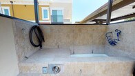 This outdoor pet wash station is a purpose-built amenity designed to make pet grooming convenient and accessible. The station features a fully tiled basin constructed with warm beige stone-look tiles that line both the walls and the basin surface, creating a clean and cohesive aesthetic. The raised washing basin is sized to accommodate pets comfortably, with built-in tethering hooks along the tiled surround to help secure animals during washing. A coiled black hose is mounted to the wall for easy access and storage, while stainless steel fixtures, including faucet controls and a drain panel, are integrated into the tile surround for a polished, durable finish. The station is situated in a partially covered outdoor area, providing shelter from direct sunlight while maintaining an open-air environment. The overhead structure allows for ample natural light and ventilation. Surrounding architecture is visible in the background, indicating this amenity is part of a larger residential community. The overall design balances functionality with an attractive finish, making it a practical and well-appointed space for pet owners.