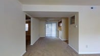 This single-floor space presents a straightforward, functional layout with a calm, neutral aesthetic throughout. The main living area is generously proportioned, finished with wall-to-wall beige carpeting that adds warmth underfoot. Smooth white ceilings are complemented by clean white baseboards, and the walls are painted in a soft, neutral tone that contributes to a bright, airy feel.

A ceiling fan is centrally positioned in the living area, enhancing both airflow and practicality. A full-height sliding glass door fitted with vertical blinds serves as a prominent feature, allowing natural light to filter into the space and providing access to the outdoors. Adjacent to the living area, a pass-through opening connects to what appears to be a kitchen space, where warm wood-toned cabinetry is visible overhead, offering practical storage.

A structural soffit detail defines the transition between areas, adding subtle architectural interest. Additional rooms are accessible via a doorway visible from the main living space, suggesting a connected corridor or hallway. Overall, the space maintains a clean, move-in-ready condition with a simple, unfussy design sensibility.