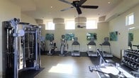 This single-floor fitness center is thoughtfully designed to provide a comprehensive workout environment. The space features a generous open layout with a combination of hardwood-style and rubberized flooring, clearly delineating cardio and strength training zones. A lineup of treadmills and elliptical machines lines the main wall, offering ample options for cardiovascular exercise. A multi-station cable strength machine is positioned along the side, expanding the range of available workouts. Ceiling fans are mounted overhead, promoting air circulation and comfort during intense exercise sessions. Natural light pours in through a series of elevated windows positioned high along the walls, creating a bright and energizing atmosphere while maintaining privacy. The walls are painted in a light neutral tone and are accented with colorful abstract artwork, adding a vibrant and motivating aesthetic to the space. Recessed lighting supplements the natural light, ensuring the area remains well-lit at all times. The overall ambiance is clean, functional, and inviting, making it a well-rounded facility suited for a variety of fitness routines.