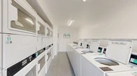 This shared laundry facility is part of a two-story residential complex constructed with classic tan brick exteriors and open-air walkways. The laundry room itself is a compact, single-floor space designed for efficiency and practicality. Inside, the room is finished in clean white throughout — walls, ceiling, and cabinetry — creating a bright and sanitary environment. Fluorescent strip lighting runs along the ceiling, ensuring the space is well-illuminated at all hours. A row of top-load washing machines lines one side of the room, while stacked washer-dryer units are mounted along the opposite wall, maximizing the available floor space. Instructional signage is posted for user reference, and durable speckled tile flooring completes the utilitarian interior. The surrounding property features generously sized surface parking lots with clearly marked spaces, making laundry trips convenient for residents. Mature trees dot the grounds, providing natural shade and a green backdrop to the complex. A covered bike rack and paved walkways are also present on the property, supporting everyday resident needs. The overall setting reflects a well-maintained, community-oriented residential property with practical shared amenities.