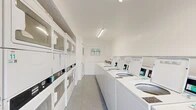 This shared laundry facility is part of a two-story residential complex constructed with classic tan brick exteriors and open-air walkways. The laundry room itself is a compact, single-floor space designed for efficiency and practicality. Inside, the room is finished in clean white throughout — walls, ceiling, and cabinetry — creating a bright and sanitary environment. Fluorescent strip lighting runs along the ceiling, ensuring the space is well-illuminated at all hours. A row of top-load washing machines lines one side of the room, while stacked washer-dryer units are mounted along the opposite wall, maximizing the available floor space. Instructional signage is posted for user reference, and durable speckled tile flooring completes the utilitarian interior. The surrounding property features generously sized surface parking lots with clearly marked spaces, making laundry trips convenient for residents. Mature trees dot the grounds, providing natural shade and a green backdrop to the complex. A covered bike rack and paved walkways are also present on the property, supporting everyday resident needs. The overall setting reflects a well-maintained, community-oriented residential property with practical shared amenities.