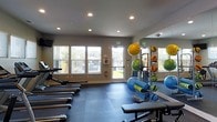 This single-floor fitness center is designed to support a wide range of workout routines in a clean, functional environment. The space is well-lit by a combination of recessed ceiling lights and multiple windows that allow natural light to fill the room, creating an open and energizing atmosphere. Large windows and a glass-paneled door face the outdoors, offering views of the surrounding grounds and enhancing the sense of space within the facility. The flooring features interlocking foam mats, providing a comfortable and safe surface for floor-based exercises and stretching. A row of treadmills lines one side of the room, offering ample cardio options for users. On the opposite side, a full-length mirror spans the wall, reflecting the space and making the room feel even more expansive while allowing users to monitor their form during workouts. A dedicated storage rack holds an assortment of medicine balls and exercise balls in various sizes, alongside rolled yoga mats, catering to both strength and flexibility training. A wall-mounted television is also present, adding entertainment options during workouts. Safety features, including a fire extinguisher and an emergency exit sign, are clearly visible. Overall, the fitness center provides a practical and inviting setting for a comprehensive exercise experience.