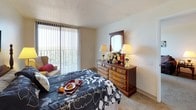Located at 4032 N Main St. in Dayton, Ohio, this single-floor unit offers a well-organized and functional layout designed for comfortable everyday living. The bedroom is a standout feature, with plush carpeting, generous natural light streaming through large floor-to-ceiling windows dressed with vertical blinds, and direct access to a private balcony overlooking lush outdoor greenery. The room comfortably accommodates full bedroom furnishings, including a four-poster bed and a multi-drawer dresser with mirror. The kitchen is efficiently designed with warm oak cabinetry lining both walls, providing ample storage and counter space. It is equipped with a full-size refrigerator and range, making it well-suited for everyday meal preparation. The bathroom features a wood-framed vanity mirror, an oak under-sink cabinet, and a shower-tub combination, all finished with clean, neutral tile flooring. The living area is carpeted and open, flowing naturally into a dedicated dining space near the entryway. A sliding glass door in the living room provides additional balcony access and fills the space with natural light and outdoor views. Crown molding and textured ceilings are consistent throughout, lending a cohesive and tidy finish to the overall interior.