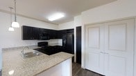 This single-floor residence offers a thoughtfully designed layout centered around a striking kitchen that serves as the heart of the home. The kitchen is outfitted with rich, dark cabinetry that provides ample storage space, complemented by light granite countertops that create a bold visual contrast. A peninsula with a built-in sink adds both additional workspace and a natural division between the kitchen and the adjoining living areas. Black appliances, including a range, built-in microwave, and a French door refrigerator, lend a cohesive and modern aesthetic throughout the space. A dedicated wine rack built into the upper cabinetry adds a refined touch for entertaining. The flooring features dark hardwood-style planks that run throughout, grounding the space with warmth and durability. Crisp white walls and flush overhead lighting keep the interior bright and clean. A paneled white door leads to additional storage or an adjoining room, contributing to the practical flow of the home. Overall, the space balances contemporary style with everyday functionality, making it a comfortable and visually cohesive living environment.