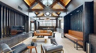 This thoughtfully designed single-floor amenity space makes an immediate impression with its dramatic vaulted ceiling, where exposed dark steel and warm timber beams intersect beneath a generous skylight that floods the interior with natural light. Light-toned hardwood flooring flows seamlessly throughout, unifying the various functional zones within the open layout.

The main lounge area is anchored by a supple cognac leather sofa paired with gray upholstered seating and a low wooden coffee table, creating an inviting gathering space. Nearby, a sleek entertainment wall provides additional relaxation options. A dedicated dining zone features a substantial dark wood table and black chairs, positioned beside a linear gas fireplace set into a gray subway tile surround. An open metal shelving unit and large industrial-style windows bring in abundant outdoor views, adding depth and brightness to the area.

The kitchenette is well-appointed with dark wood cabinetry, a matte stone countertop, an undermount sink with a professional-style faucet, and floor-to-ceiling windows overlooking the exterior. A distinctive lounge nook showcases a textured mosaic accent wall paired with chartreuse bench seating. The fitness area is visible through bold vertical metal slat partitions, with exercise equipment bathed in natural light. A dedicated bike storage room completes this versatile, design-forward space.