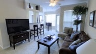 This single-floor space offers an open-concept design that seamlessly connects the living and dining areas, creating a cohesive and functional environment. The living area is anchored by plush, comfortable seating arranged around a dark wood coffee table, complemented by soft carpeting underfoot. A ceiling fan with a light fixture provides both comfort and ambient illumination.

A raised breakfast bar with dark wood barstool seating serves as a natural divider between the living space and the kitchen area, adding both style and convenience. Decorative accents, including tall vases and candle holders displayed on the bar ledge, lend a polished, curated feel to the space.

The dining area, positioned near large windows with horizontal blinds, benefits from generous natural light that brightens the entire floor plan. A pendant light fixture hangs above the dining table, adding a warm focal point to the space.

Crown molding along the upper walls adds a refined architectural detail, while the neutral color palette throughout creates a clean, inviting atmosphere. Multiple windows ensure the space remains bright throughout the day, offering an airy, open ambiance. The thoughtful layout and consistent design language make this a comfortable and visually appealing living environment.