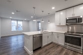 This thoughtfully designed single-floor unit offers a clean, contemporary aesthetic throughout. The heart of the space is an open-concept kitchen that seamlessly connects to the living area. The kitchen is outfitted with white shaker-style cabinetry, light quartz countertops, and a full suite of stainless steel appliances including a French door refrigerator, slide-in range, built-in microwave, and dishwasher. A generously sized peninsula island with a double undermount sink serves as both a functional workspace and a casual dining area, highlighted by a trio of polished pendant lights overhead. Recessed lighting throughout ensures consistent illumination across all spaces. Light wood-style luxury vinyl flooring runs continuously throughout the unit, adding warmth and visual cohesion. The living area is bright and open, benefiting from large windows that bring in natural light and offer views of the surrounding neighborhood. The bedrooms are well-proportioned, each featuring the same durable flooring and neutral gray wall tones. The unit includes two bathrooms: one featuring a sleek glass-enclosed walk-in shower, and the other equipped with a full soaking tub and shower combination with chrome fixtures. A connecting hallway leads to additional rooms, completing this modern and functional floor plan.