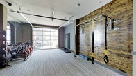 This single-level fitness studio is thoughtfully designed to support a wide range of workout routines in a clean, contemporary setting. The space is anchored by a striking reclaimed wood accent wall outfitted with a yellow-framed functional training rig, complete with TRP suspension straps, pull-up bars, and resistance accessories — providing a versatile station for bodyweight and functional strength training. The open floor plan is finished with light-toned, low-pile carpeting that offers a comfortable surface underfoot while maintaining a polished aesthetic. One of the most notable architectural features is the full-width glass garage door along the exterior wall, which opens the space to the outdoors and allows abundant natural light to pour in, creating an airy and energizing atmosphere. The high ceiling, accented by ceiling fans and recessed lighting, enhances the sense of openness and ensures proper airflow during intense workouts. A mirrored wall panel provides visual feedback for form and technique, while additional fitness equipment — including stationary bikes — lines the perimeter. The overall ambiance is modern and motivating, blending industrial design elements with functional fitness infrastructure.