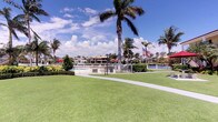 This vibrant outdoor space embodies the essence of Florida waterfront living, offering a beautifully maintained environment that seamlessly blends lush landscaping with stunning aquatic surroundings. The grounds are defined by an expansive, immaculately manicured lawn that provides an inviting open area for leisure and recreation. Tall, graceful palm trees are scattered throughout, lending the space a distinctly tropical character and offering natural shade beneath bright, sunny skies. A gently curving walkway winds through the grounds, guiding visitors toward the waterfront area where a fenced pool facility is situated. The pool zone is framed by well-kept hedges and ornamental plantings, adding a sense of privacy and polish. Beyond the pool, the shimmering water opens up to picturesque views of neighboring waterfront homes, lush foliage, and a prominently displayed American flag that adds a patriotic accent to the scenery. The overall ambiance is one of relaxed, resort-style outdoor living, with the combination of open green space, water views, and tropical landscaping creating a welcoming and visually dynamic environment. Whether used for casual strolling, poolside relaxation, or simply enjoying the scenery, this space captures the spirit of warm-weather coastal living at its finest.
