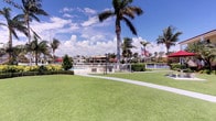This vibrant outdoor space embodies the essence of Florida waterfront living, offering a beautifully maintained environment that seamlessly blends lush landscaping with stunning aquatic surroundings. The grounds are defined by an expansive, immaculately manicured lawn that provides an inviting open area for leisure and recreation. Tall, graceful palm trees are scattered throughout, lending the space a distinctly tropical character and offering natural shade beneath bright, sunny skies. A gently curving walkway winds through the grounds, guiding visitors toward the waterfront area where a fenced pool facility is situated. The pool zone is framed by well-kept hedges and ornamental plantings, adding a sense of privacy and polish. Beyond the pool, the shimmering water opens up to picturesque views of neighboring waterfront homes, lush foliage, and a prominently displayed American flag that adds a patriotic accent to the scenery. The overall ambiance is one of relaxed, resort-style outdoor living, with the combination of open green space, water views, and tropical landscaping creating a welcoming and visually dynamic environment. Whether used for casual strolling, poolside relaxation, or simply enjoying the scenery, this space captures the spirit of warm-weather coastal living at its finest.