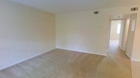 This single-floor space offers a straightforward and functional layout designed with simplicity in mind. The interior is finished in soft, neutral tones throughout, with smooth walls painted in a warm off-white that contributes to a bright and open atmosphere. Wall-to-wall beige carpeting covers the floors, providing a comfortable and cohesive look that complements the overall color palette. The main living area is generously proportioned, offering ample room for a variety of furniture arrangements. Crisp white baseboards frame the perimeter of the room, adding a clean, finished appearance. A ceiling-mounted HVAC vent ensures climate control is well integrated into the space. Electrical outlets and switch plates are conveniently positioned throughout, reflecting practical design choices. The open, uncluttered layout makes this space highly adaptable, whether configured as a living room, bedroom, or multi-purpose area. Natural light filters into the room, enhancing the warmth of the neutral finishes. A doorway visible along one wall provides access to additional areas of the space, suggesting a connected and flowing floor plan. Overall, this space presents a clean, move-in ready environment with a calm and understated aesthetic.