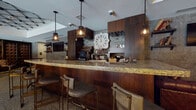 Located on East Ninth Street in Cleveland, Ohio, this single-floor bar and lounge space presents a thoughtfully curated interior that blends industrial and upscale design elements throughout. The layout is expansive, accommodating multiple distinct seating zones that cater to various guest experiences within one cohesive environment.

At the heart of the space is a well-appointed bar featuring a polished granite countertop, rich dark wood cabinetry, and draft beer taps. Pendant lighting with cage-style fixtures hangs above the bar, casting a warm glow over the leather-upholstered bar stools. A large decorative clock serves as a focal point behind the bar, flanked by organized glassware displays and mirrored shelving.

The lounge areas are furnished with tufted leather Chesterfield sofas and armchairs arranged around low coffee tables, anchored by geometric-patterned area rugs. Terrazzo flooring extends throughout the space, adding a classic textural quality to the floors. Dark stone-clad accent walls contribute to the moody, intimate atmosphere, while large windows along one side allow natural light and views of the surrounding Cleveland streetscape.

A grid-style track lighting system overhead and a statement chandelier provide layered illumination. Additional seating nooks with high-top tables and bookcases with decorative accessories add depth and character to the overall space.