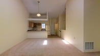 This two-story space is defined by its striking vaulted ceilings, which lend an airy, open quality to the main living area. The layout follows an open-concept design, where the living room flows seamlessly into the kitchen, creating a cohesive and functional environment. Soft, neutral-toned carpeting covers the living area floor, contributing to a warm and understated ambiance throughout.

The kitchen is positioned on a slightly elevated platform and features wood-toned cabinetry, a white refrigerator, a range with overhead ventilation, and dark countertops. A peninsula-style breakfast bar separates the kitchen from the living space, providing additional counter space and a casual dining option. A pendant light hangs above this transition area, adding a functional focal point to the open layout.

Natural light filters into the kitchen through a window above the sink, brightening the space. A door visible at the rear of the kitchen leads to an adjacent area, likely a utility or storage room. The second floor is accessible via a staircase visible toward the right side of the space, with the upper level overlooking the main living area below, reinforcing the home's open vertical design.