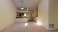 This two-story space is defined by its striking vaulted ceilings, which lend an airy, open quality to the main living area. The layout follows an open-concept design, where the living room flows seamlessly into the kitchen, creating a cohesive and functional environment. Soft, neutral-toned carpeting covers the living area floor, contributing to a warm and understated ambiance throughout.

The kitchen is positioned on a slightly elevated platform and features wood-toned cabinetry, a white refrigerator, a range with overhead ventilation, and dark countertops. A peninsula-style breakfast bar separates the kitchen from the living space, providing additional counter space and a casual dining option. A pendant light hangs above this transition area, adding a functional focal point to the open layout.

Natural light filters into the kitchen through a window above the sink, brightening the space. A door visible at the rear of the kitchen leads to an adjacent area, likely a utility or storage room. The second floor is accessible via a staircase visible toward the right side of the space, with the upper level overlooking the main living area below, reinforcing the home's open vertical design.