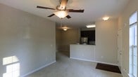 This single-floor residence features an open-concept layout that connects the living area to the kitchen, creating a seamless and functional flow throughout the space. The living area is finished with neutral-toned carpeting and warm beige walls, lending a clean and understated aesthetic. A ceiling fan with an integrated light fixture is centrally mounted, providing both air circulation and ambient lighting. The kitchen is defined by a raised breakfast bar pass-through countertop featuring granite-style surfaces, offering a practical divide between cooking and living spaces while maintaining an open feel. Dark espresso-toned cabinetry lines the kitchen walls, complementing the stainless steel refrigerator that adds a modern touch. Recessed and flush-mount lighting provides consistent illumination throughout the kitchen area. White baseboards frame the perimeter of the space, adding a crisp contrast to the neutral walls and carpet. The overall design emphasizes practicality and simplicity, with an open floor plan that maximizes the sense of space and encourages ease of movement between functional areas.