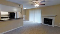 This single-story space features an open, functional layout that connects the living area and kitchen in a practical design. The living room is carpeted and centered around a classic fireplace with a tiled surround, providing a warm focal point. A ceiling fan with an integrated light fixture is installed overhead, contributing to both comfort and ambient lighting. Large sliding glass doors dressed with vertical blinds span one wall, allowing natural light to fill the space while offering access to the outdoors. The kitchen is partially open to the living area via a raised pass-through counter finished with a dark countertop, allowing for easy interaction between the two spaces. A wall-mounted mirror in the kitchen area adds depth and reflects additional light throughout. The warm yellow-toned walls, white baseboards, and neutral carpet create a cohesive and inviting interior palette. Overhead ventilation is provided by a ceiling vent in the living area, supporting efficient climate control. Overall, the space offers a well-balanced blend of comfort and practicality.