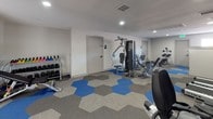 This fitness room offers a functional and well-organized workout space designed to accommodate a range of exercise needs. The room features clean white walls and recessed lighting that keeps the space bright and practical. A standout design element is the geometric hexagonal carpet, rendered in a striking combination of blue, grey, and neutral tones, which adds visual interest to the floor while providing a durable workout surface.

The room is outfitted with a multi-station cable and weight machine positioned centrally, suitable for a wide variety of strength training exercises. An adjustable weight bench and additional resistance machines are arranged along one side, offering options for both upper and lower body workouts. A dedicated free weight area includes a well-stocked dumbbell rack with weights ranging across multiple sizes, along with a flat bench for supplementary exercises.

Two interior doors provide access to additional areas, likely a restroom or storage. The overall layout is efficient and uncluttered, allowing ample space to move between equipment. The space presents a clean, modern aesthetic suited for regular fitness use.