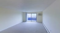 This single-floor space offers a clean, functional layout centered around a spacious main living area. The room is finished with neutral wall-to-wall carpeting and crisp white walls, lending it a bright and open feel. A large sliding glass door serves as the focal point of the living area, flooding the space with natural light and providing direct access to a private balcony. The balcony overlooks surrounding buildings and mature trees, offering a pleasant outdoor retreat. A wall-mounted air conditioning unit ensures year-round climate comfort, while a baseboard heating system runs along the perimeter of the room, providing an additional source of warmth during cooler months. The overall design is straightforward and versatile, offering a blank canvas well-suited for a variety of furnishing arrangements. The clean lines, abundant natural light, and practical amenities make this a highly functional living space.