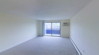 This single-floor space offers a clean, functional layout centered around a spacious main living area. The room is finished with neutral wall-to-wall carpeting and crisp white walls, lending it a bright and open feel. A large sliding glass door serves as the focal point of the living area, flooding the space with natural light and providing direct access to a private balcony. The balcony overlooks surrounding buildings and mature trees, offering a pleasant outdoor retreat. A wall-mounted air conditioning unit ensures year-round climate comfort, while a baseboard heating system runs along the perimeter of the room, providing an additional source of warmth during cooler months. The overall design is straightforward and versatile, offering a blank canvas well-suited for a variety of furnishing arrangements. The clean lines, abundant natural light, and practical amenities make this a highly functional living space.