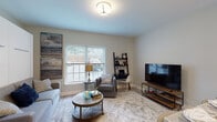 Located at 502 Howe Ave in Shelton, Connecticut, this thoughtfully designed single-floor unit offers an efficient and stylish layout that maximizes every square foot. The open-concept living area is bathed in natural light through large windows fitted with white blinds, creating a bright and welcoming atmosphere. Light wood-tone flooring flows consistently throughout the space, complementing the neutral warm-beige wall tones.

A standout feature of the living area is the built-in Murphy bed system, seamlessly integrated behind a clean white cabinet facade, paired with a sofa and flanked by built-in storage drawers — an ideal space-saving solution. The living room also accommodates a seating arrangement with accent artwork and a dedicated media area.

The kitchen is a highlight of the unit, showcasing crisp white shaker cabinetry with crown molding, sparkling granite countertops, and a full suite of stainless steel appliances including a refrigerator, electric range, and over-the-range microwave. A peninsula with bar seating for three provides a casual dining option beneath pendant lighting.

The bathroom features marble-look tile flooring, a gray shaker-style vanity with a white countertop, a framed mirror, and elegant wall sconce lighting, delivering a polished, contemporary finish throughout.