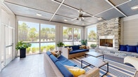 This screened lounge is a well-appointed single-level space designed to blend comfort with an open, airy atmosphere. Large screen-panel walls on the pool-facing side flood the interior with natural light and offer unobstructed views of a sparkling pool, palm trees, and lush landscaping beyond. The coffered metal ceiling, accented by a prominent industrial-style ceiling fan and recessed lighting, adds a refined coastal character overhead. Durable tile flooring runs throughout, complementing the space's indoor-outdoor aesthetic. The seating area centers around a generous wicker sectional and matching sofa, both dressed in bold navy cushions with gold and patterned accent pillows, arranged over a navy-and-gold striped area rug. A sleek coffee table anchors the grouping. On the opposite wall, a floor-to-ceiling stacked stone fireplace serves as a dramatic focal point, flanked by a built-in bench with blue cushions and accented by a sculptural yellow lounge chair. A wall-mounted flat-screen television is positioned nearby for entertaining. Horizontal shiplap-style wall paneling in crisp white runs throughout, lending a clean, contemporary coastal feel. Potted tropical plants add warmth and greenery, while a glass door connects to an adjacent covered corridor, enhancing the overall flow of the space.