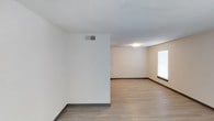 This single-floor space offers a straightforward and practical layout designed for comfortable everyday living. The interior features light wood-style laminate flooring throughout, which lends warmth and continuity to the open areas. Neutral, light-toned walls create a bright and airy atmosphere, while a textured ceiling adds a subtle layer of visual detail overhead. Dark baseboards provide a clean contrast against the pale walls and flooring, giving the space a polished, finished look. A ceiling-mounted light fixture provides consistent overhead illumination, complementing the natural light that filters in through a window fitted with horizontal blinds. The HVAC vent positioned on the upper wall indicates a central air system, contributing to year-round comfort. The open floor plan allows for flexible furniture arrangements and accommodates a range of living configurations. The overall ambiance is clean, minimal, and move-in ready, making it a versatile space well-suited for those seeking a simple, functional environment with a neutral palette as a backdrop for personalization.